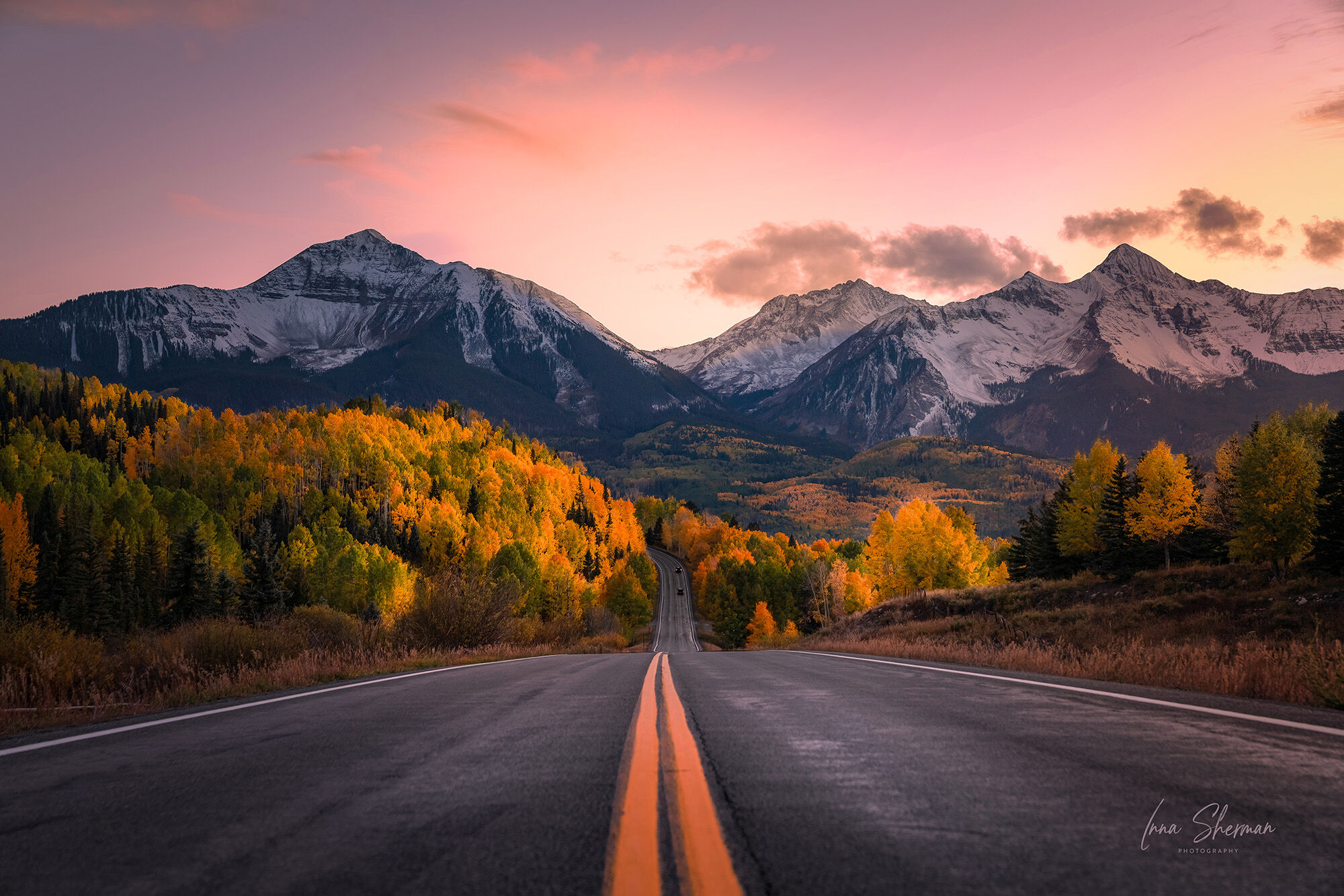 A mountain road in Colorado at sunset, lined with golden aspens and leading toward snow-dusted peaks under a pink sky. Photo by Inna Sherman.