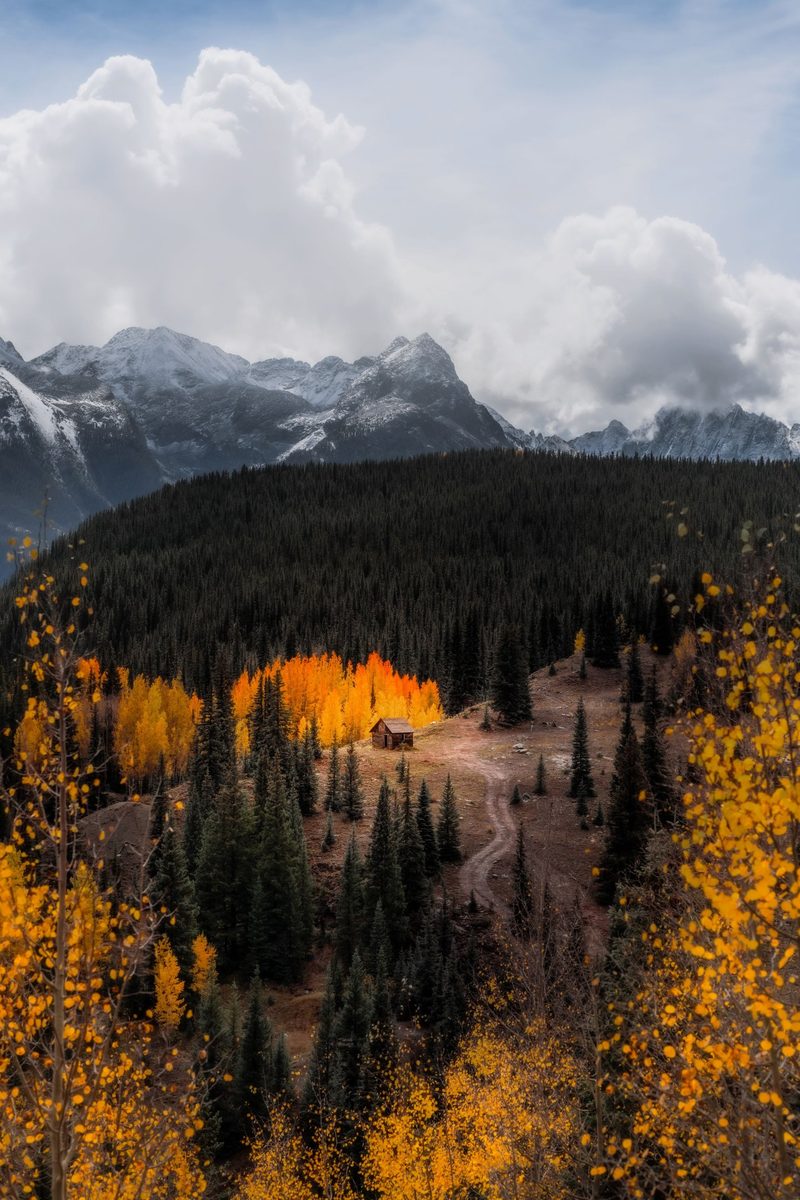 A small cabin lit by glowing orange aspens below snowy peaks and a dark forest