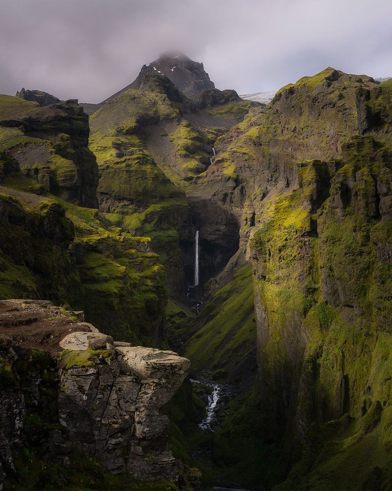 A thin waterfall deep in a green moss-covered gorge beneath a cloud-capped peak