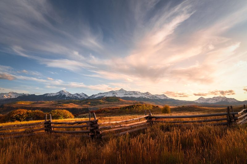 A split-rail fence in golden grass before snow-covered peaks under sweeping clouds