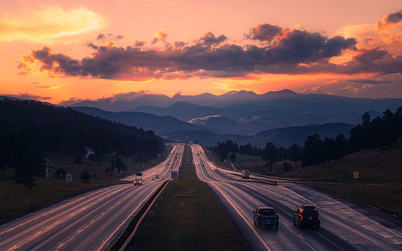A highway heading west into layered blue mountains under a blazing orange sunset