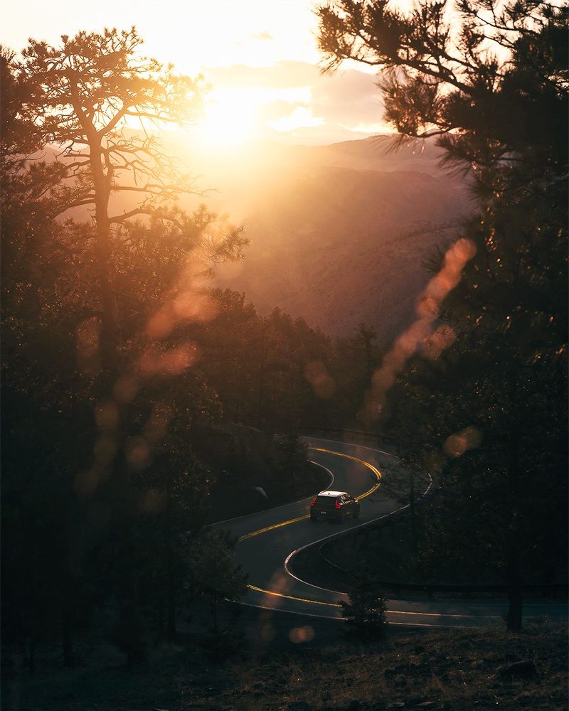 A car winding down a serpentine mountain road through pines at sunset