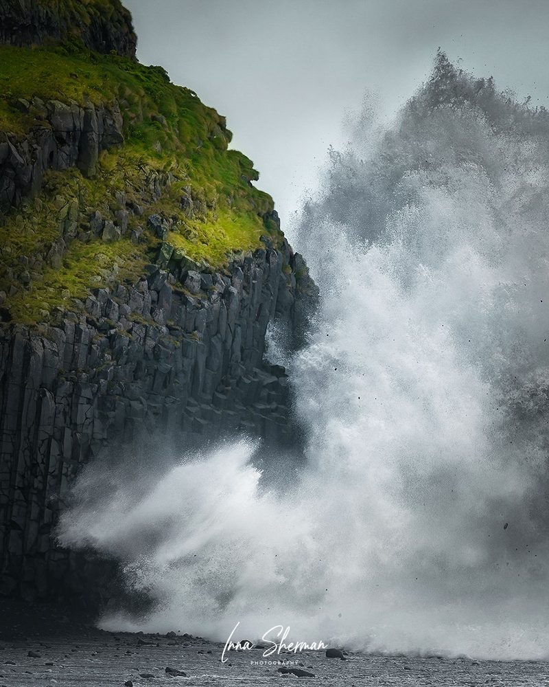 A wave exploding against a basalt-column sea cliff topped with green grass