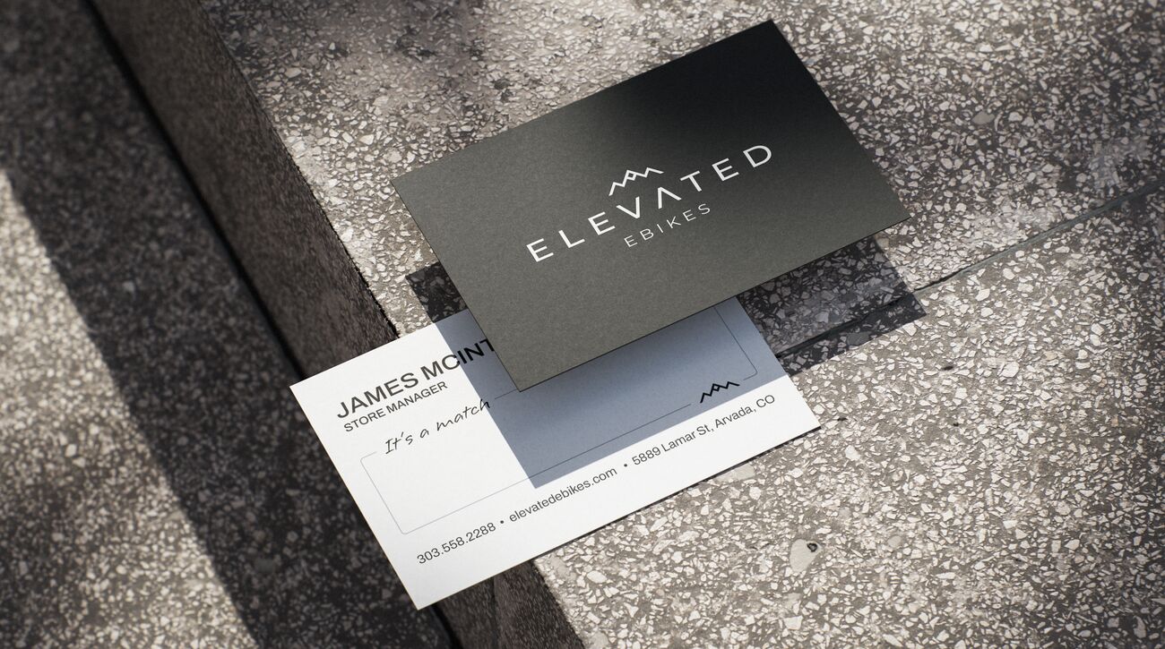 Elevated Ebikes business cards on a terrazzo step: a black front with the ELEVATED EBIKES logo, and a white back with the owner’s name and title, a handwritten note reading It’s a match, the shop’s phone, website and Lamar St address, and the peak mark