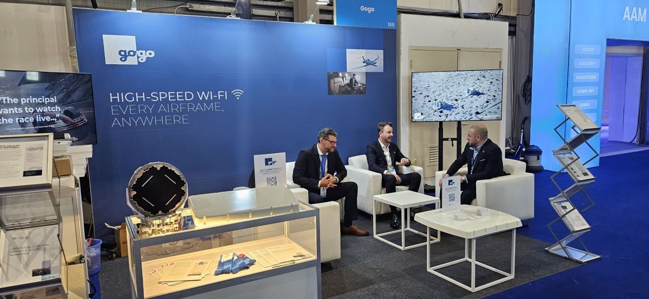Gogo booth at the Dubai Airshow: a navy wall reading High-speed Wi-Fi, every airframe, anywhere, an antenna in a glass case, and three people talking in white lounge chairs
