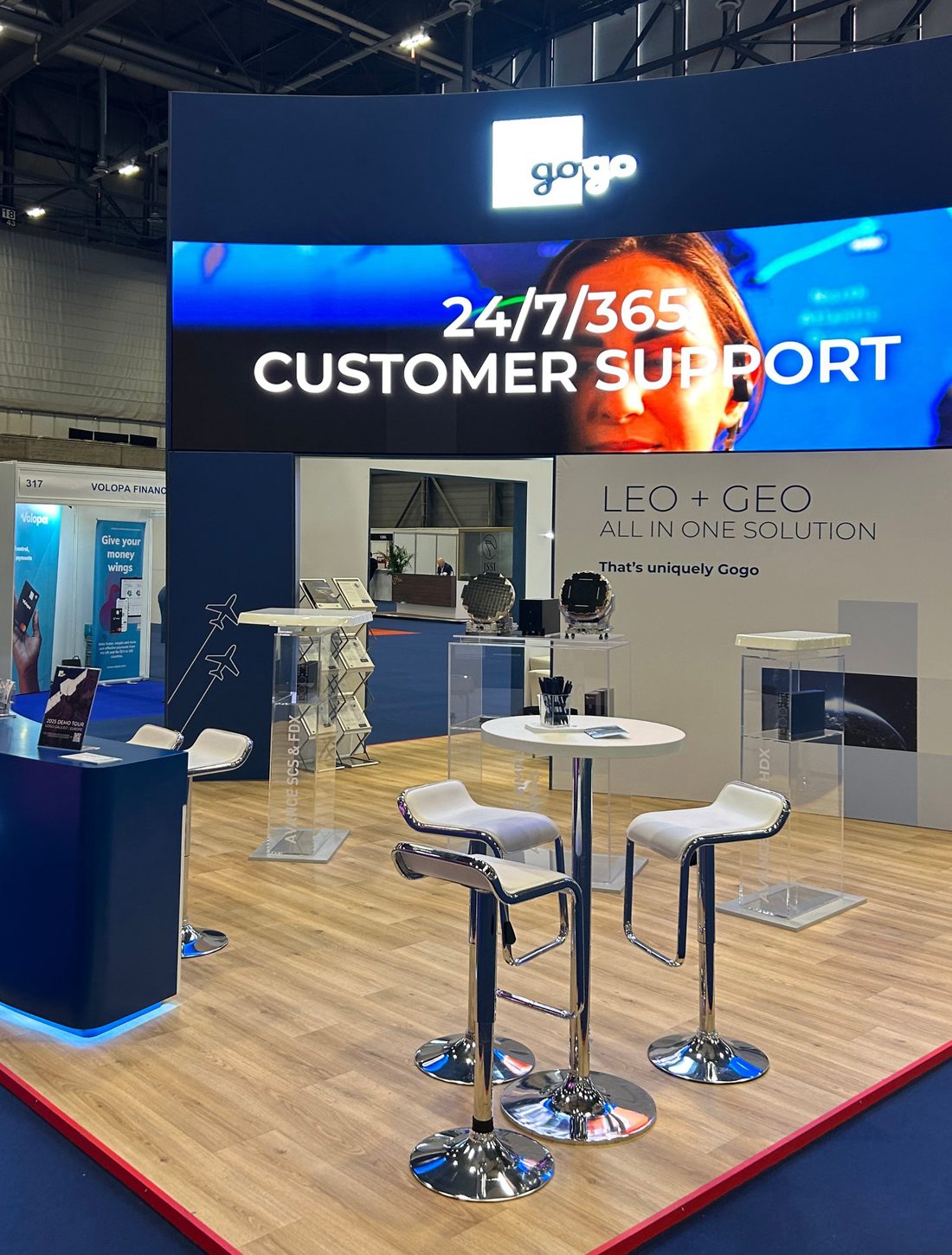 EBACE 2025 Gogo booth with an LED wall reading 24/7/365 Customer Support, a navy reception counter, white stools and antennas in acrylic cases