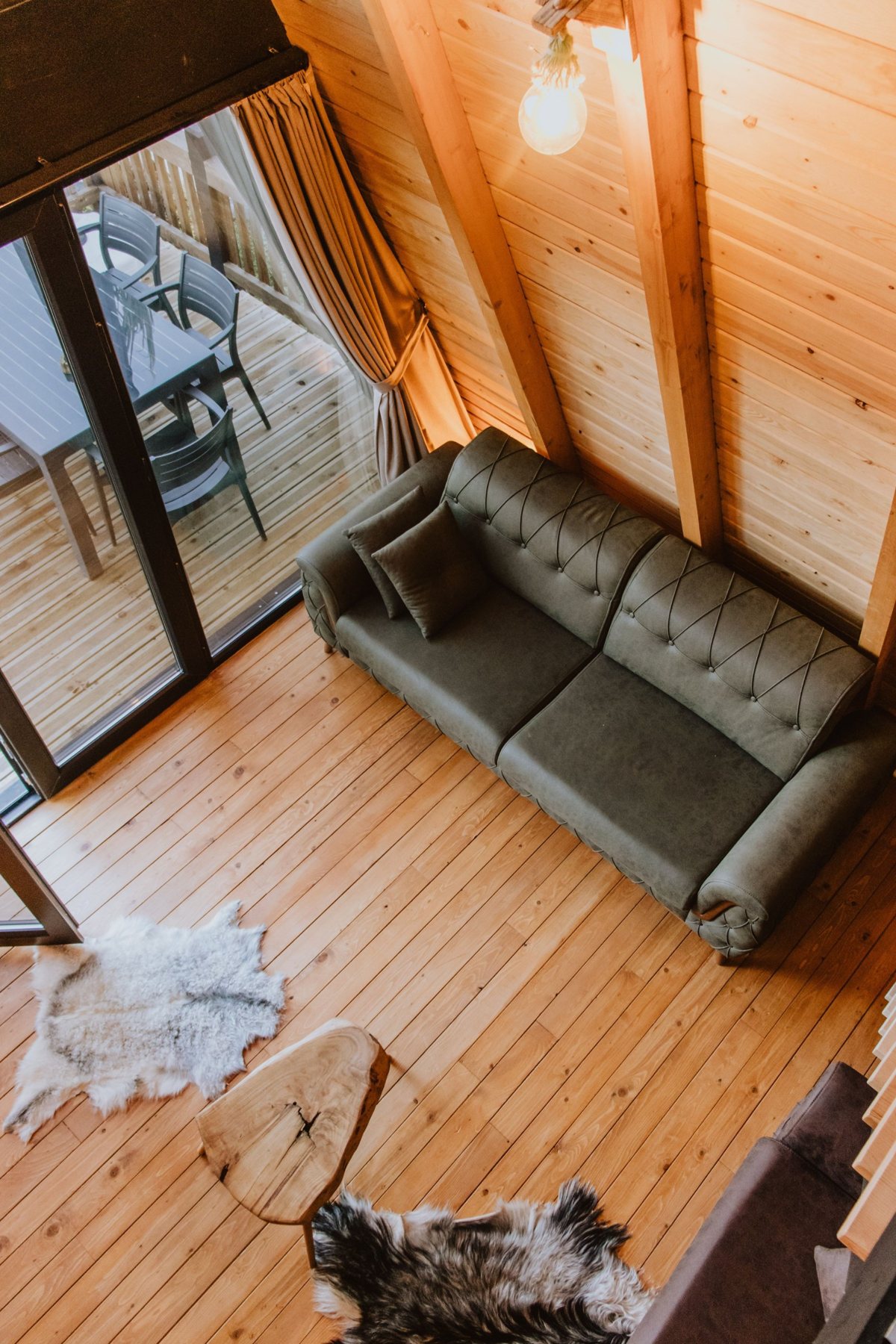 Looking down into a warm wood-paneled cabin room with a green sofa, sheepskin rugs and a glass door to a deck