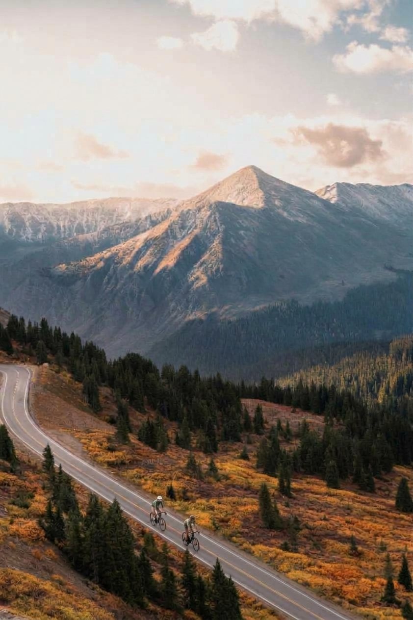 Two riders small on a paved mountain road beneath a snow-dusted peak in warm evening light, Cottonwood Pass